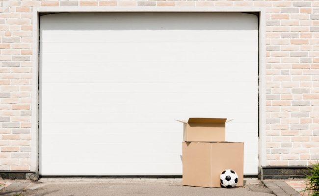 Two cardboard boxes, one open and one closed, are placed in front of a white garage door. A soccer ball rests on the ground next to them. The garage backdrop features light-colored bricks.