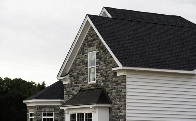 A modern house featuring a dark shingled roof, stone facade on the front gable, and white siding. Two windows are visible on the side of the house. Trees are in the background under a cloudy sky.