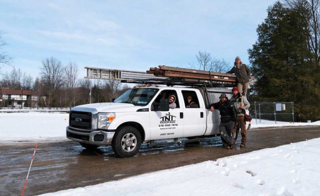 A white pickup truck with ladders on top is parked on a snowy road. Five people are around the truck, with two standing on the passenger side step and one sitting on a ladder. Trees and houses are visible in the background.