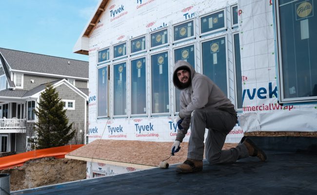 A person in a hoodie and jeans is working on a house under construction, kneeling on a roof covered with black material. The house has multiple windows and is wrapped in Tyvek. Another house and a tree are visible in the background.
