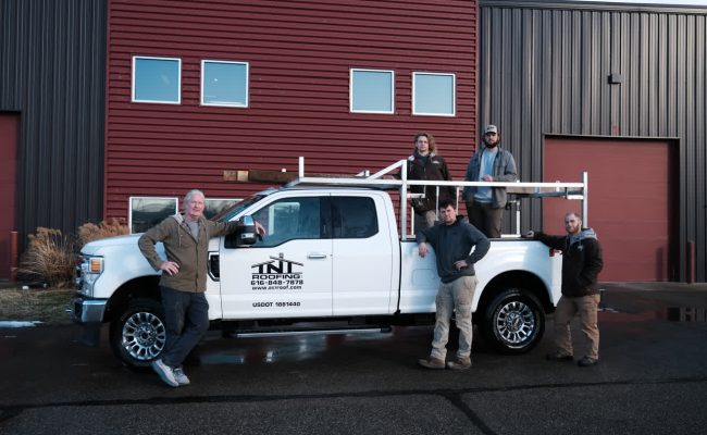 Five people are posing around a white pickup truck with a TNC Roofing logo, parked in front of a red industrial building with two garage doors and windows. The sky is clear, and the asphalt is wet.