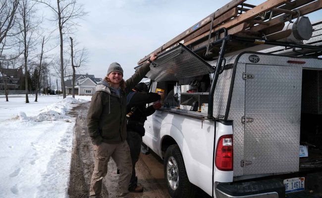 A person in winter clothing stands smiling beside a white utility truck with an open side compartment. Another person is inside, reaching into the compartment. The ground is snowy, and several bare trees are in the background.