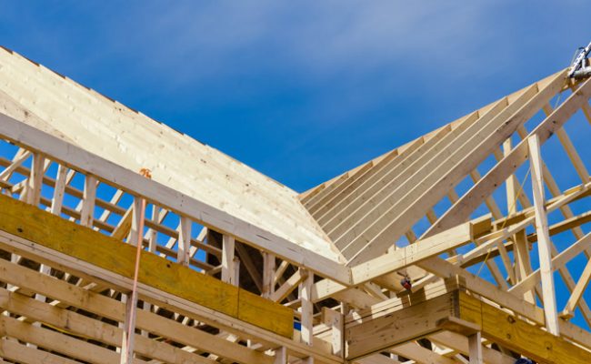 Wooden framework of a house under construction against a clear blue sky. The image shows the skeletal structure with exposed beams and trusses, highlighting the progress in the building process.